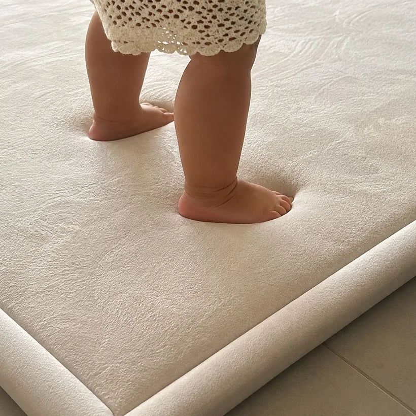 Baby standing on a soft beige Misk Mat with feet sinking into the 35mm memory foam cushioning, showing the plush texture and raised mat edge on tiled flooring.