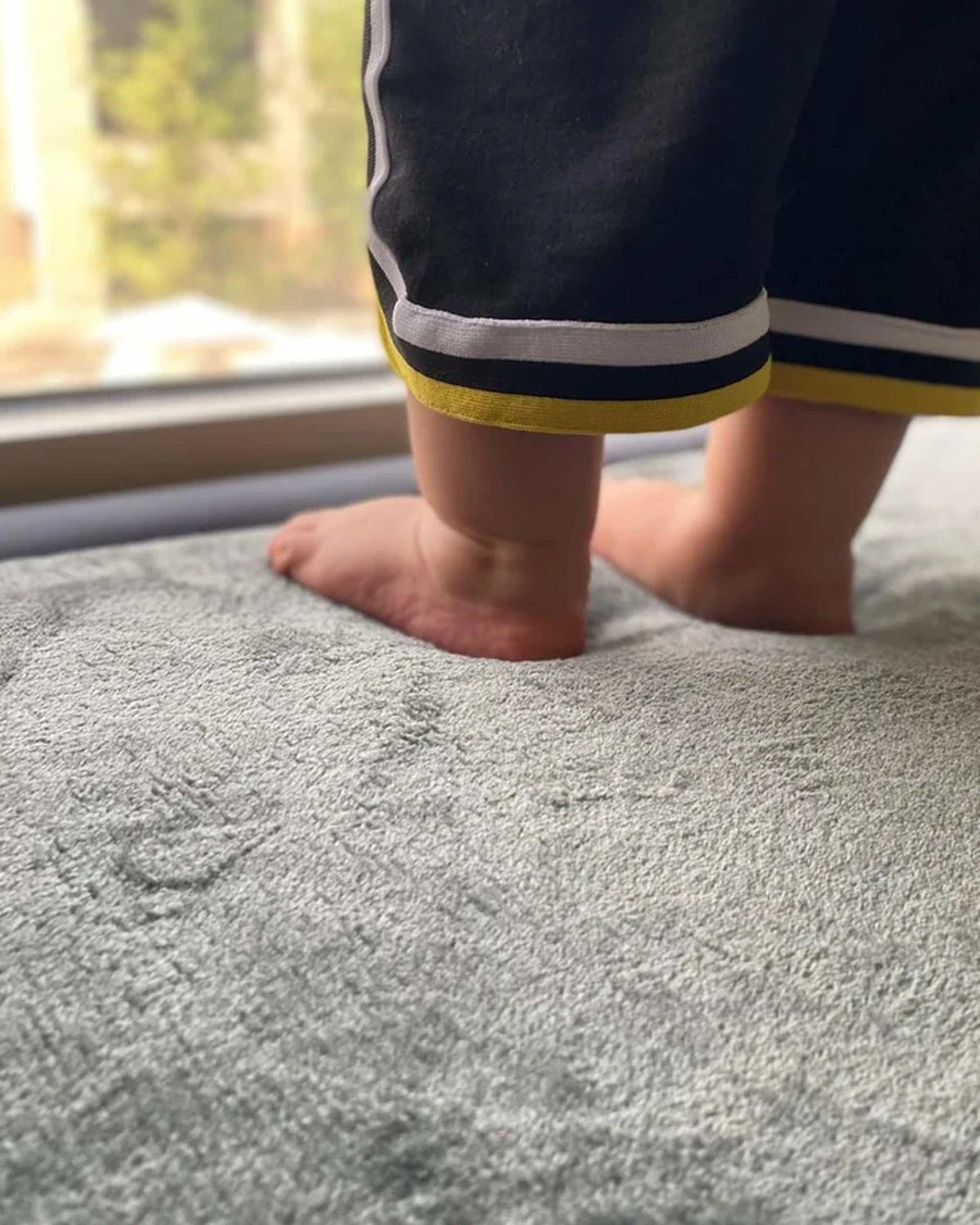 Close-up of a toddler's feet standing on a Dark Grey Misk Mat 35mm memory foam mat by a sunlit window.