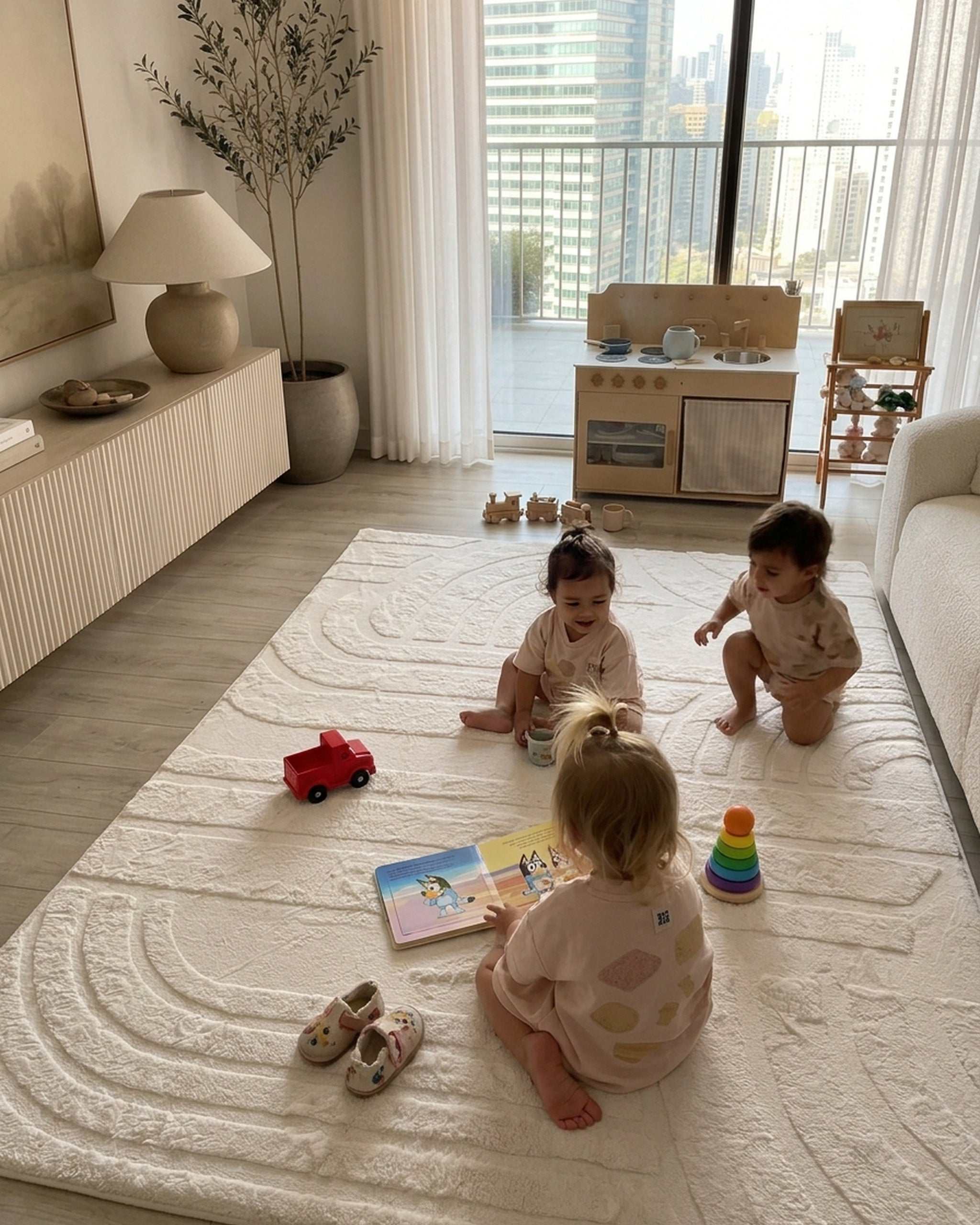 Three toddlers playing with wooden toys on a Vanilla Cream Misk Mat memory foam mat in a modern living room with a play kitchen and city skyline view.