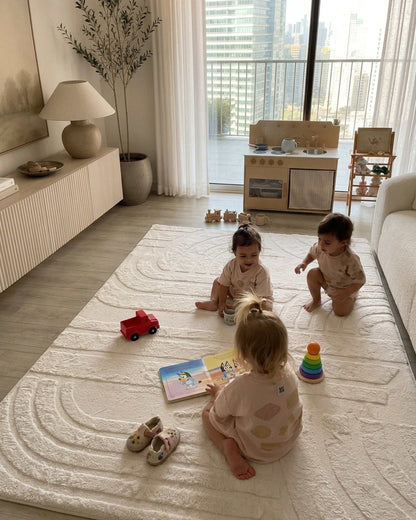 Three toddlers playing with wooden toys on a Vanilla Cream Misk Mat memory foam mat in a modern living room with a play kitchen and city skyline view.