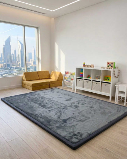 Dark Grey Misk Mat 35mm memory foam mat in a bright kids' playroom with a mustard sofa, toy shelf and city skyline view.