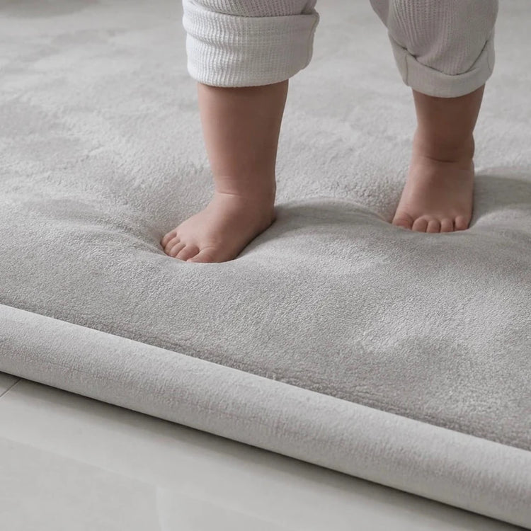 Close-up of toddler feet sinking into a soft light grey Misk Mat memory foam play mat, showing thick plush cushioning and the mat edge on a tiled floor.