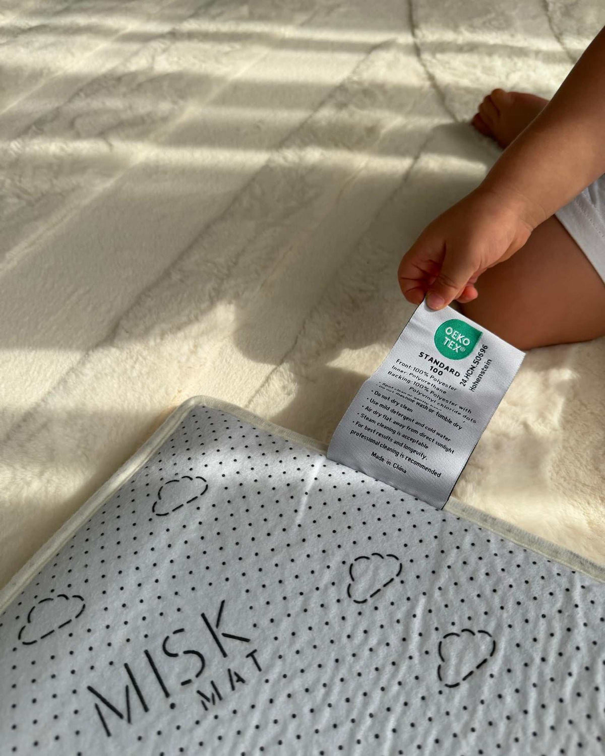 Close-up of the Misk Mat OEKO-TEX certified label sewn onto the mat backing, with a child's hand reaching for the tag.