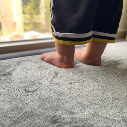 Toddler feet sinking into a soft dark grey Misk Mat memory foam play mat, showing plush cushioning and a cozy family-friendly surface near a window.